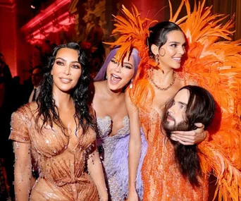 A group of Met Gala attendees in beaded and feathered outfits, one holding a prop head with a realistic face.