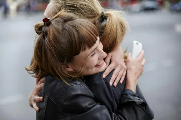 Two people happily hugging on a street, one holding a phone, both smiling warmly.