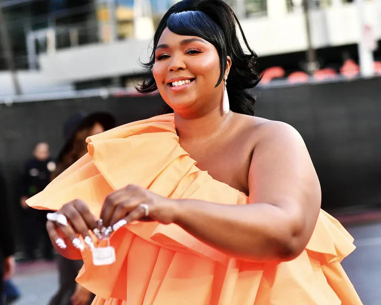 A woman in an orange ruffled dress smiles while holding a small purse, outdoors with blurred people in the background.