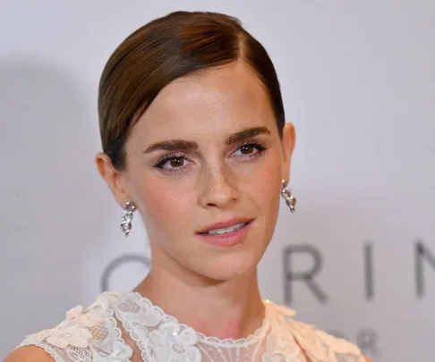 A person with short brown hair wearing a white lace top and diamond earrings at an event, with a neutral expression.