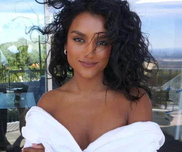 Person with curly hair enjoying a breezy day on a balcony, wrapped in a white robe, with a serene view in the background.