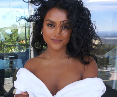 Person with curly hair enjoying a breezy day on a balcony, wrapped in a white robe, with a serene view in the background.