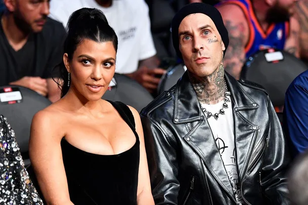 Kourtney Kardashian and Travis Barker sitting together at an event, wearing black outfits, with Travis in a leather jacket.