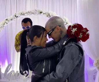 Couple in leather jackets embracing under floral arch, holding red rose bouquet, with Elvis impersonator in background.