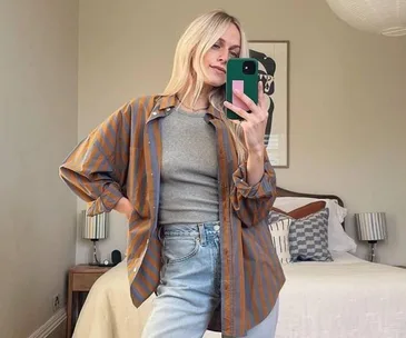 Blonde woman taking a selfie in a striped shirt over a gray top and jeans, standing in a stylish bedroom.