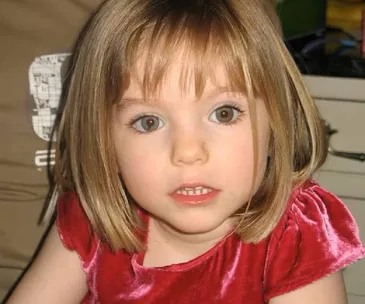 Young child with short blonde hair in a red dress, sitting indoors and looking towards the camera.