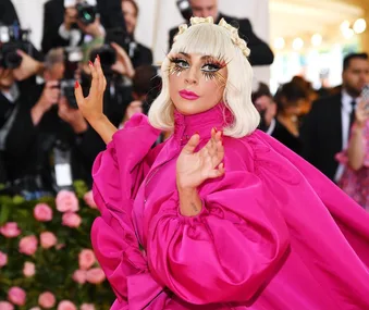 Person in a vibrant pink outfit with dramatic eye makeup, posing confidently at a high-profile event with photographers.