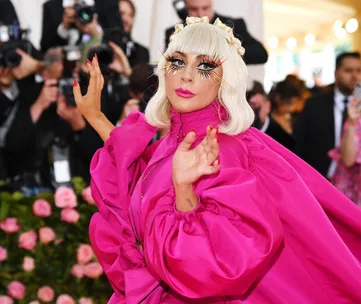 Person in a vibrant pink outfit with dramatic eye makeup, posing confidently at a high-profile event with photographers.