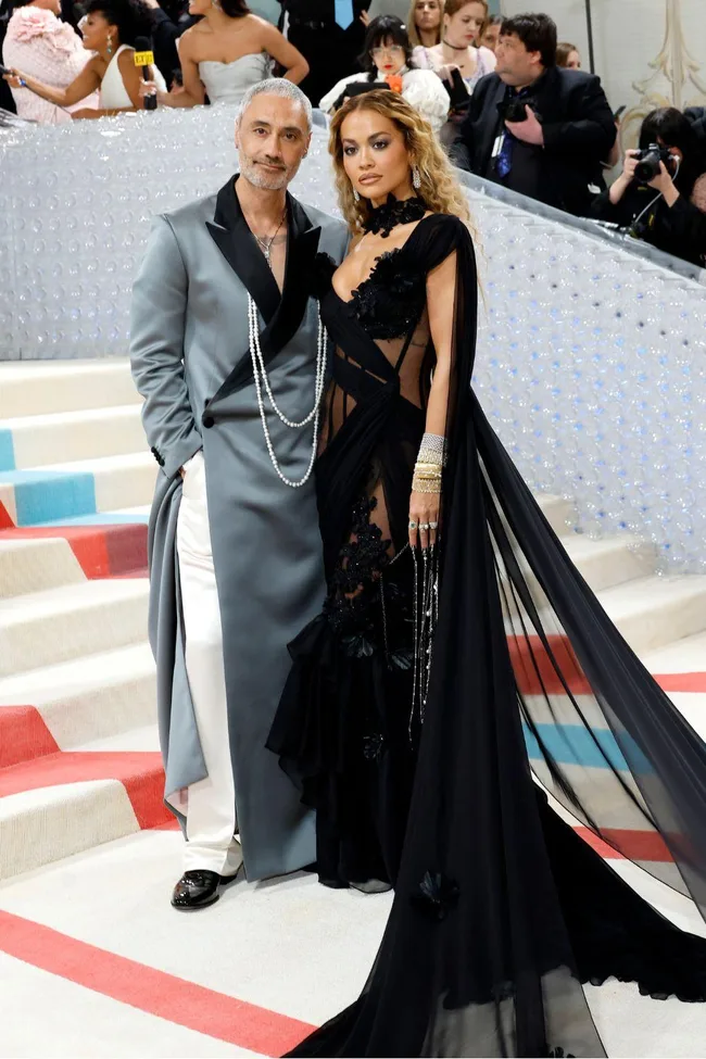 A couple poses on the Met Gala 2023 red carpet; the man wears a gray suit and the woman a sheer black gown.