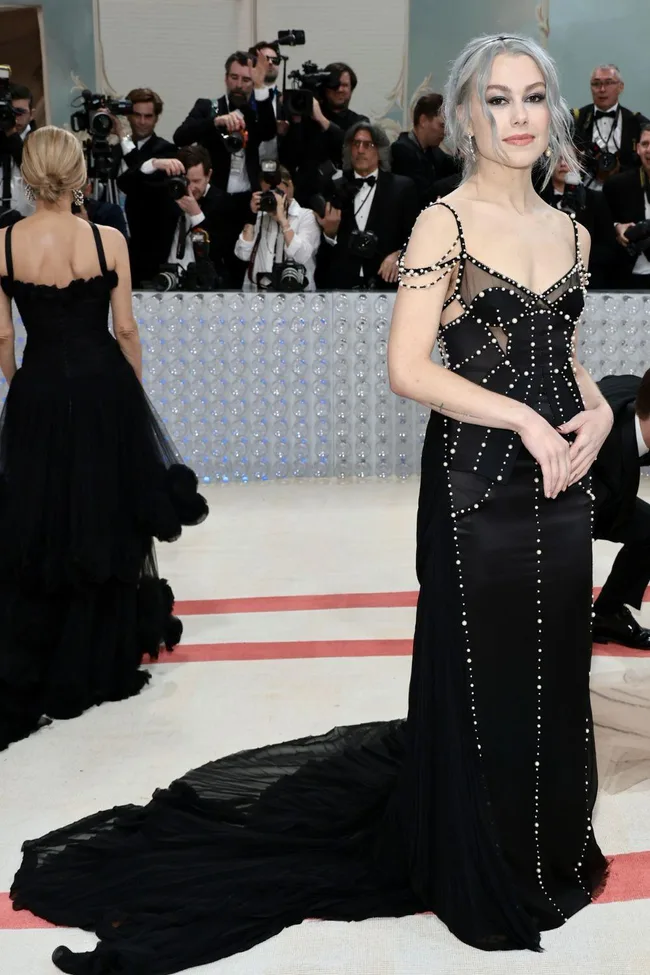 Woman in elegant black gown with pearl details and silver hair on the Met Gala 2023 red carpet, photographers in background.