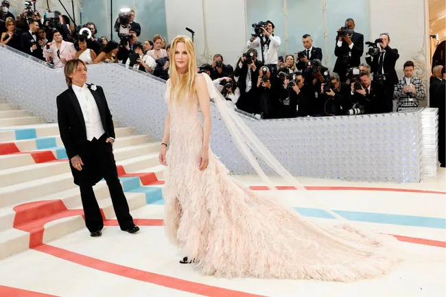 Nicole Kidman in a feathered blush gown with a train at the Met Gala 2023, accompanied by a suited man, on a red and blue carpet.