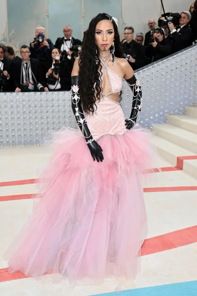 A person wearing a pink tulle gown with black floral gloves poses at the Met Gala 2023, surrounded by photographers.