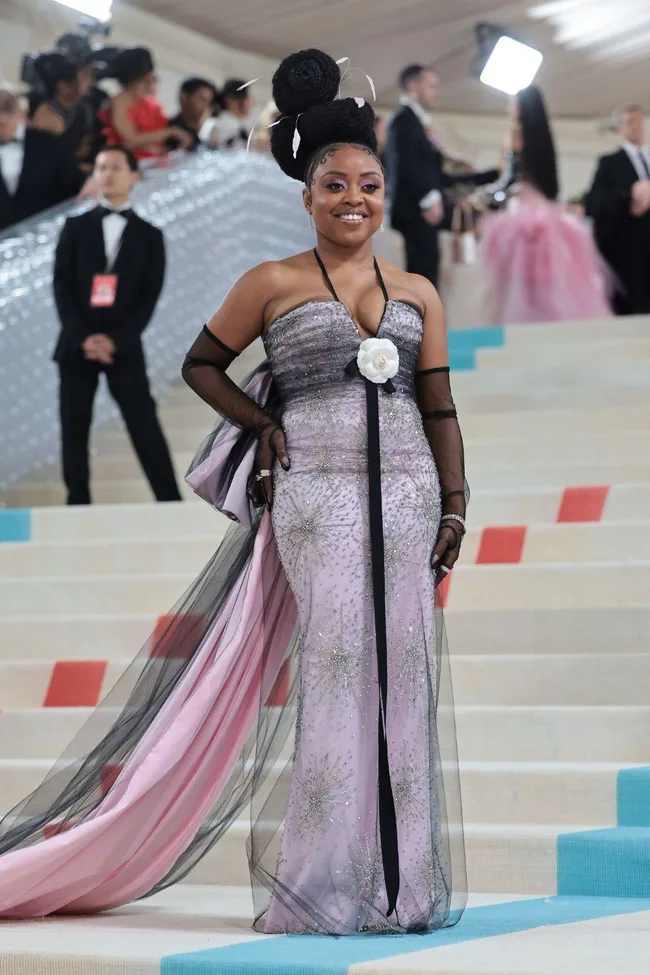 A person in a glittering lavender gown with floral details poses on the Met Gala 2023 stairs amid a crowd and photographers.