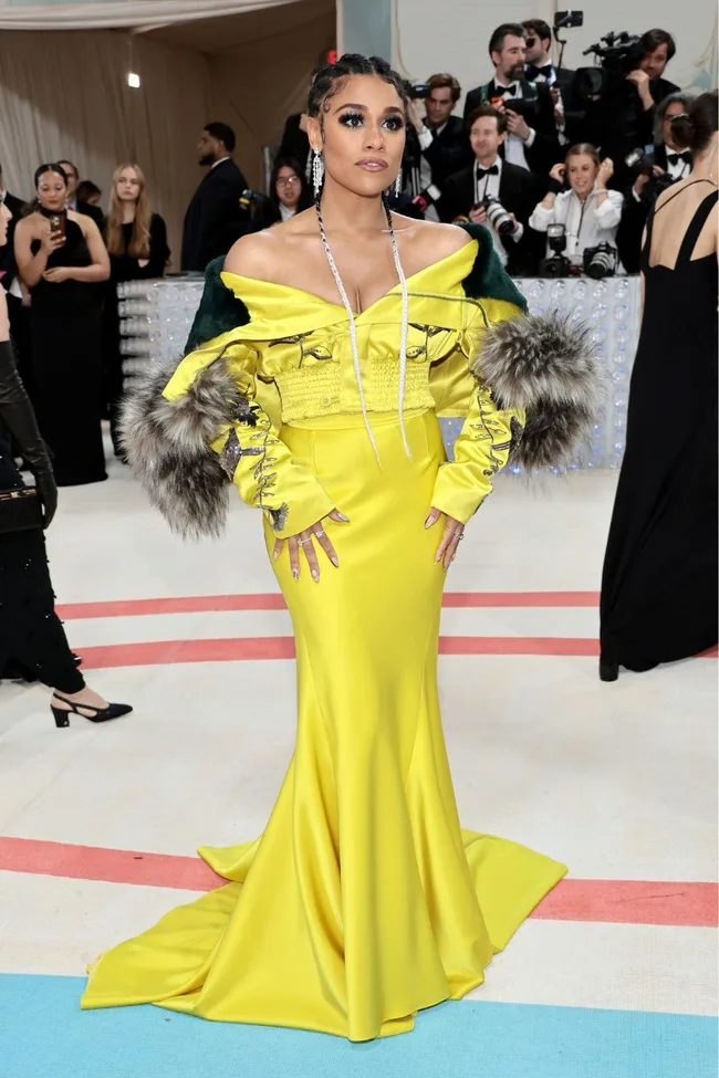 A woman in a vibrant yellow gown with fur, posing at a formal event with photographers in the background.