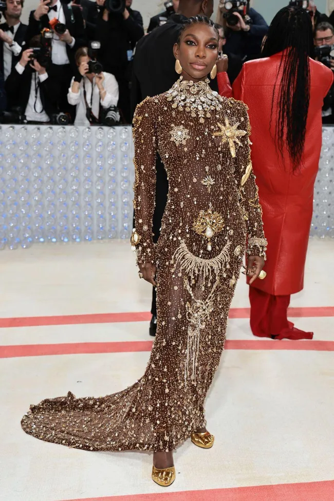 Elegant figure in a bejeweled, gold-embellished gown at Met Gala 2023, surrounded by photographers.