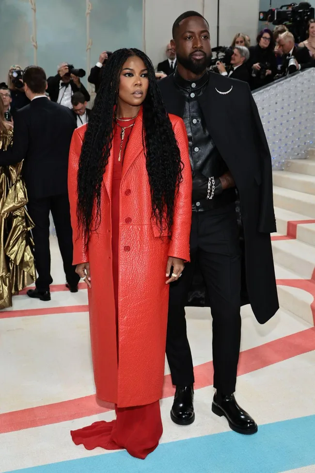 A couple poses at the Met Gala 2023; she wears a red leather coat, and he wears a black outfit with a cape.