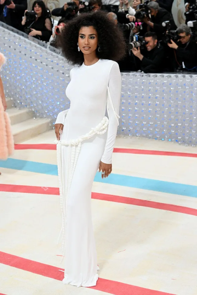 Woman in a white gown with pearls, posing at the 2023 Met Gala, surrounded by photographers on a patterned carpet.