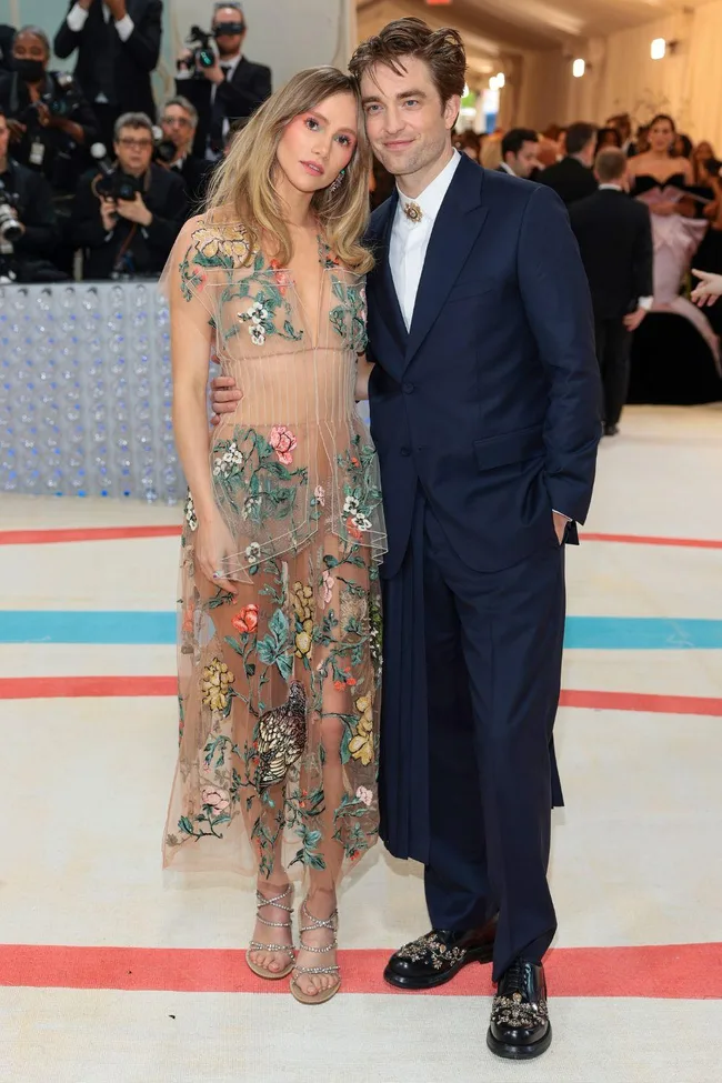 A couple poses on the Met Gala 2023 red carpet; one wears a floral sheer dress, the other a navy suit.