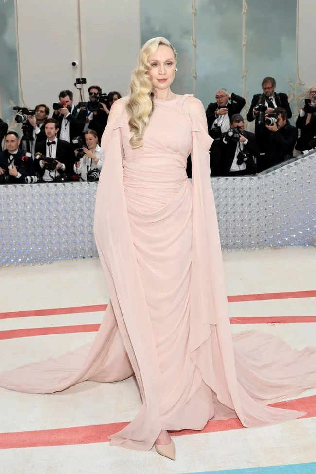 Person in a flowing pink gown poses on a red carpet while photographers capture the moment in the background.