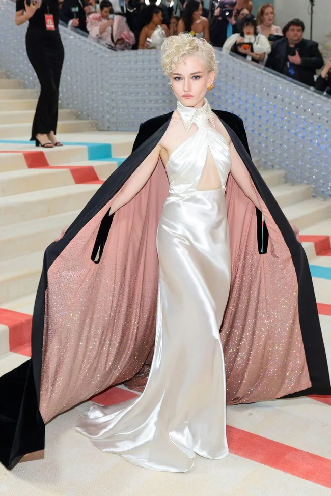 Person in a silver gown with a black cape, showcasing fashion on a staircase at a gala event.