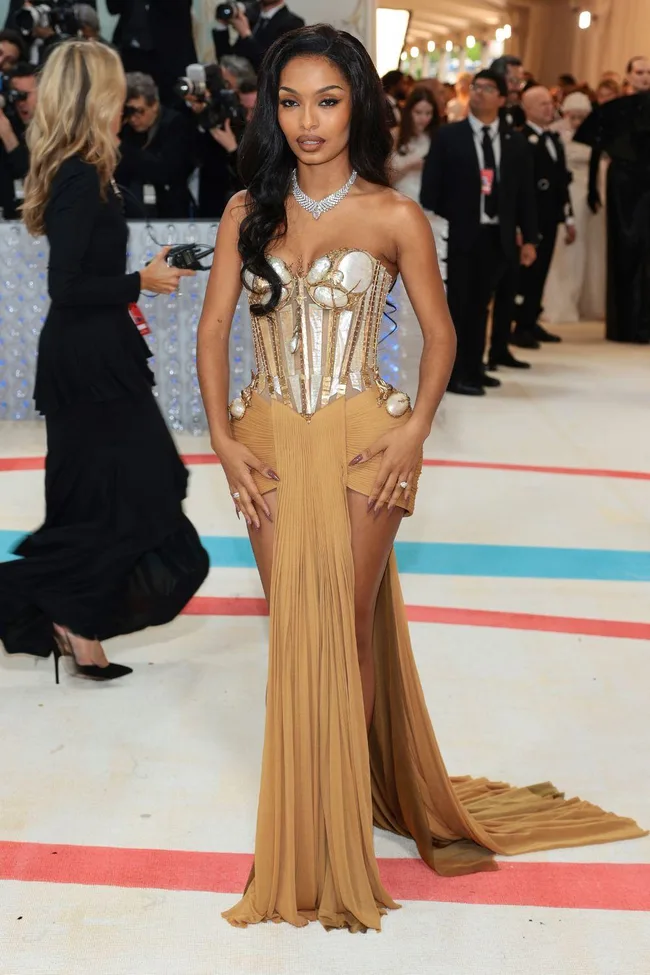 A woman in a metallic corset gown with a flowing skirt poses on the Met Gala 2023 red carpet, surrounded by photographers.