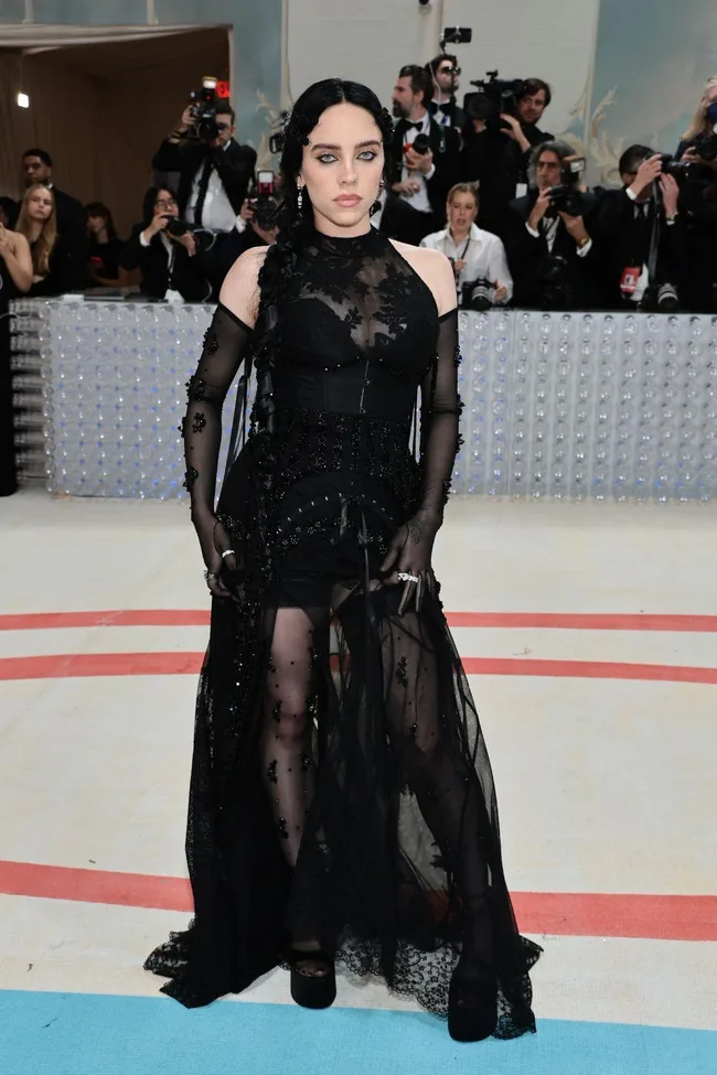 A person in an intricate black lace gown poses on the Met Gala 2023 red carpet, photographers in the background.