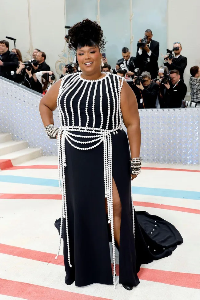 Person in a black dress with pearl adornments at Met Gala 2023, surrounded by photographers.