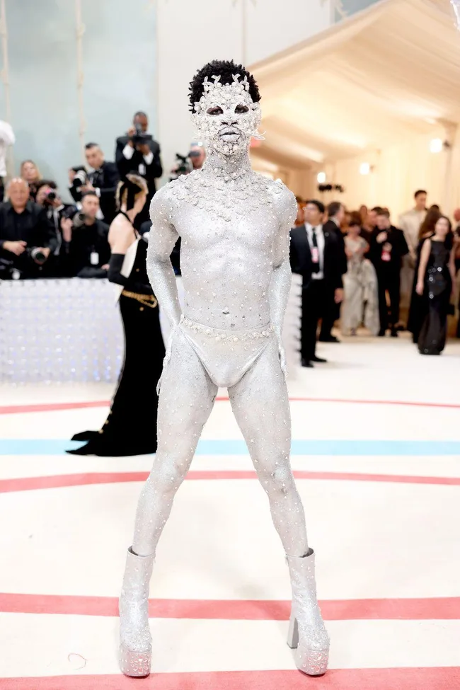 Person in a full body silver bejeweled outfit and boots, wearing an intricate headpiece and mask at the 2023 Met Gala.