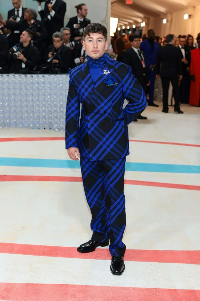 A person in a blue and black plaid suit at the Met Gala 2023, posing on a decorated carpet with photographers behind.