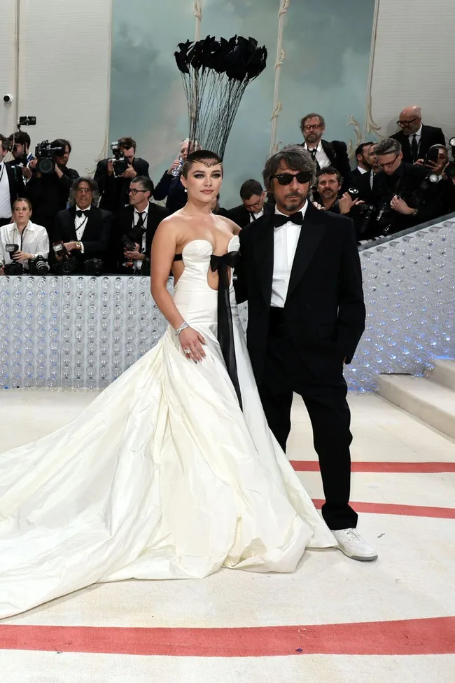 A woman in a white gown with a tall headpiece stands beside a man in a black suit at the 2023 Met Gala.