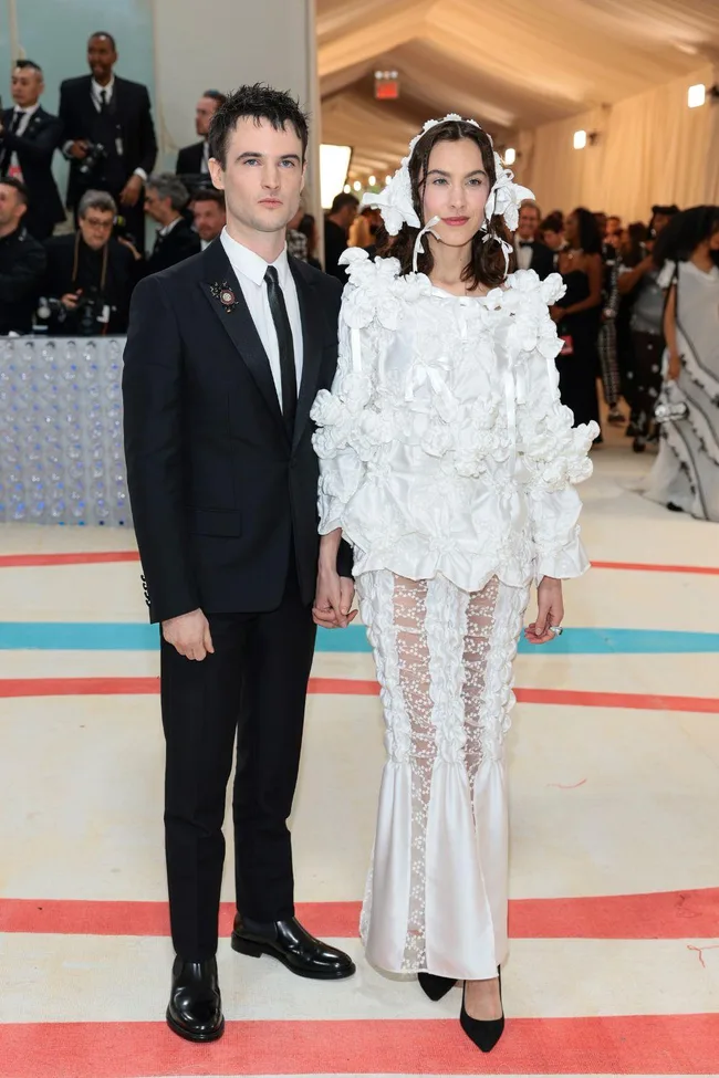 A couple poses at Met Gala 2023; man in black suit, woman in elaborate white outfit with floral details and lace.