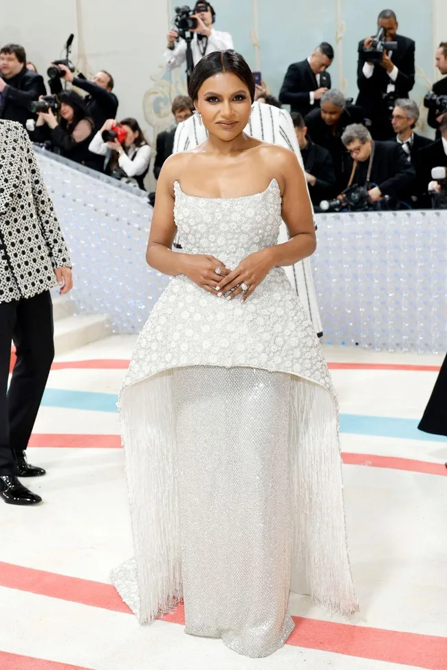 A woman in a white, strapless, embellished gown with fringe stands on a red and cream carpet, surrounded by photographers.