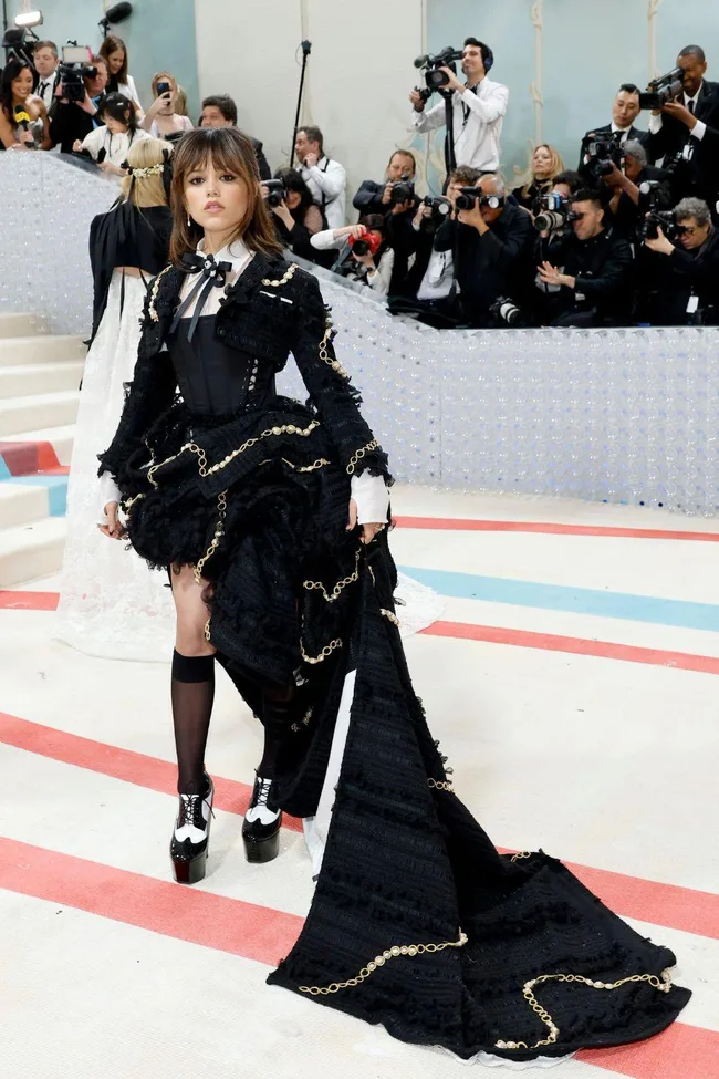 A woman at the Met Gala 2023 in a black, ruffled gown with gold chain details, surrounded by photographers.