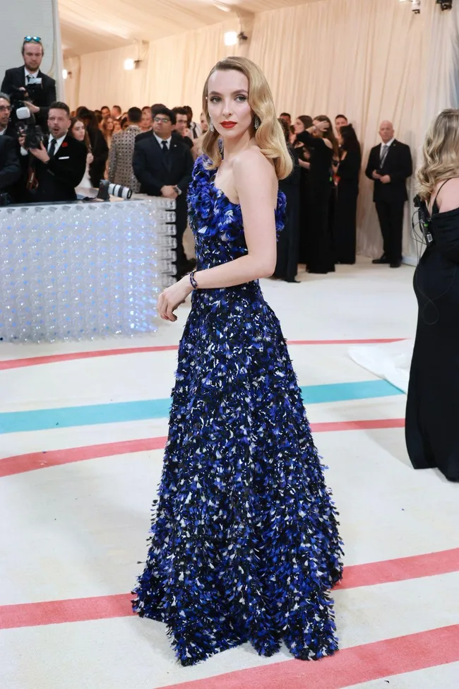 Person in a blue feathered gown at Met Gala 2023, posing on a cream-colored carpet with photographers in the background.