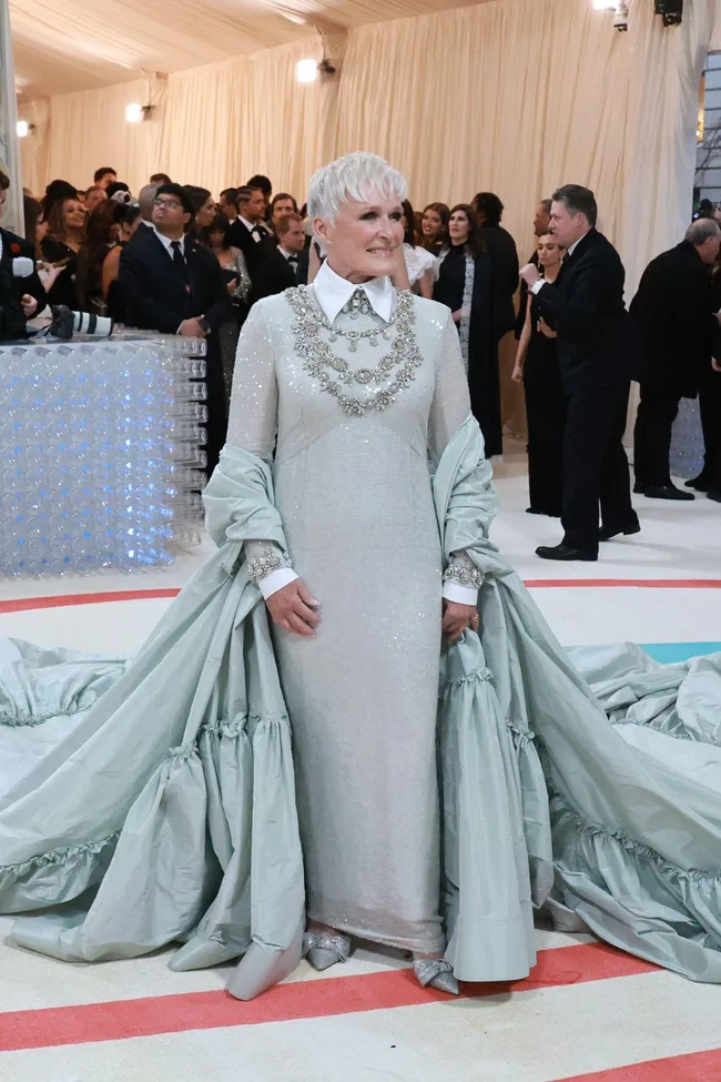 Woman in a silver gown with ornate necklace and shawl at an elegant event, surrounded by guests.