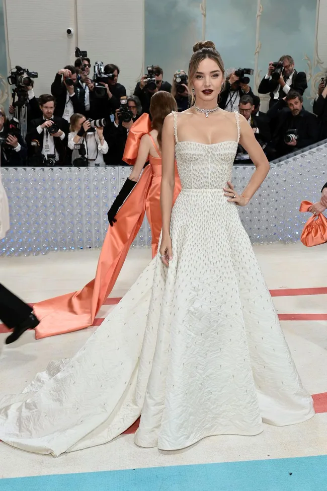 A person in an elegant white gown with a diamond necklace and hair in a bun, posing at a formal event with photographers.