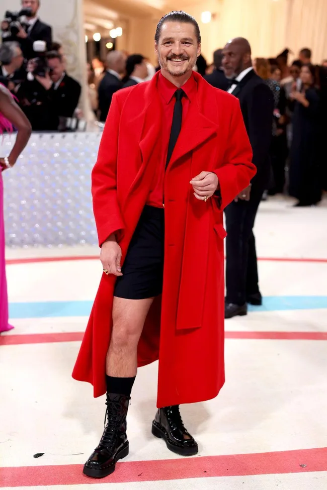 Man in a vibrant red coat and black shorts at the Met Gala 2023, smiling and posing on the red carpet.