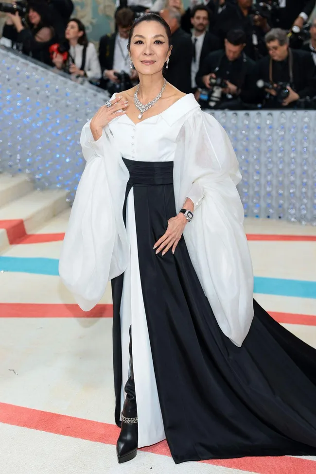 Elegant woman in a black and white gown at Met Gala 2023, posing on a red-blue carpet with photographers in background.