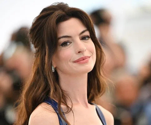 Anne Hathaway smiling at an event, wearing a blue dress and earrings, with her long hair styled down.