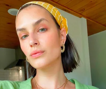 Woman with short dark hair, wearing a yellow headband, gold hoop earrings, and a green top, in a wooden-ceiling room.