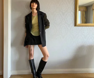 A person leans against a wall, wearing a blazer with textured sleeves, and knee-high boots in a stylish room.