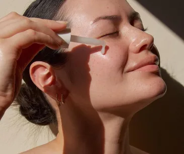 Woman applying serum on face with dropper, smiling gently in sunlight.