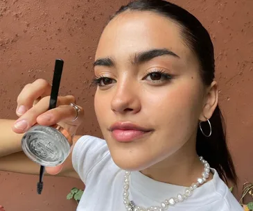 Person with sleek hair holds Anastasia Beverly Hills brow product, wearing a pearl necklace and hoop earrings.