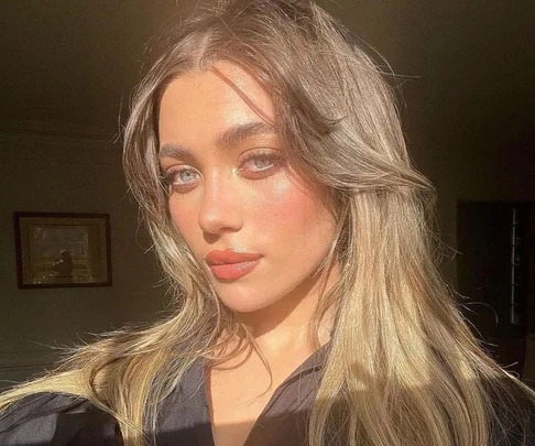 Woman with long hair and glowing skin posing in sunlight indoors.