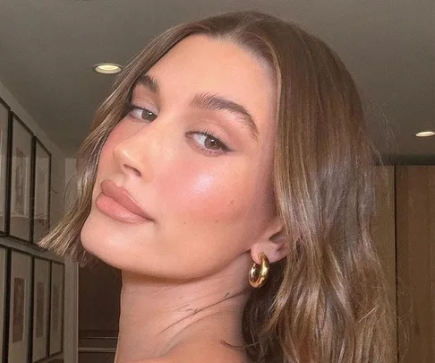 A woman with glowing skin and wavy brown hair looks over her shoulder, wearing gold hoop earrings, in a softly lit room.