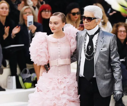 A model in a pink ruffled dress and a designer in a silver suit walk together at a fashion show, surrounded by an audience.