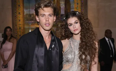 A man in a black outfit and a woman with long curly hair in a sparkly dress pose at an event.