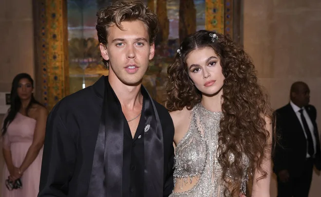 A man in a black outfit and a woman with long curly hair in a sparkly dress pose at an event.