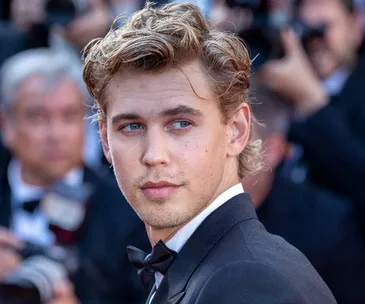 A person in a tuxedo at a formal event, gazing to the side, surrounded by a blurred crowd.