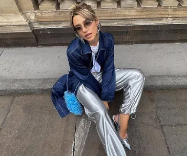 Person sitting on a city curb wearing sunglasses, a denim jacket, metallic pants, and holding a blue fluffy bag.